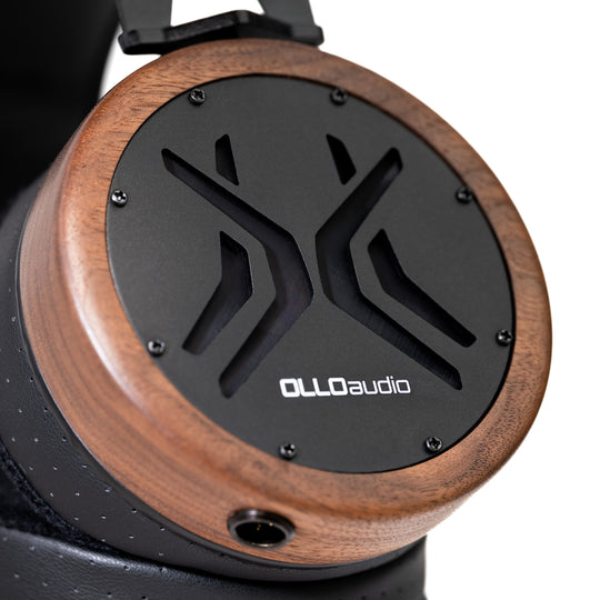 Close-up of an OLLO Audio over-ear headphone earcup featuring a black central panel with a stylized logo cutout, surrounded by a medium brown wood-grain frame and a black padded headband.