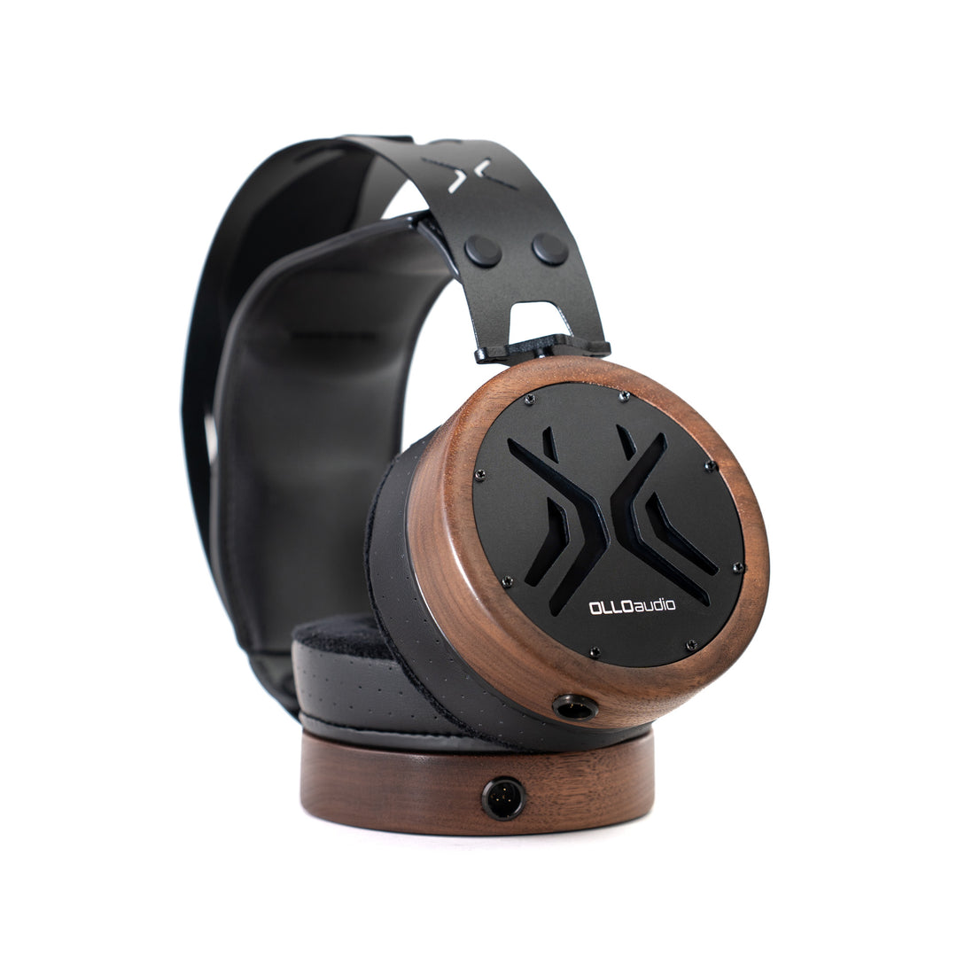 A pair of OLLO Audio over-ear headphones with a distinctive dark brown wood-grain finish on the earcups and a black headband, resting on a matching wooden display stand against a white background.